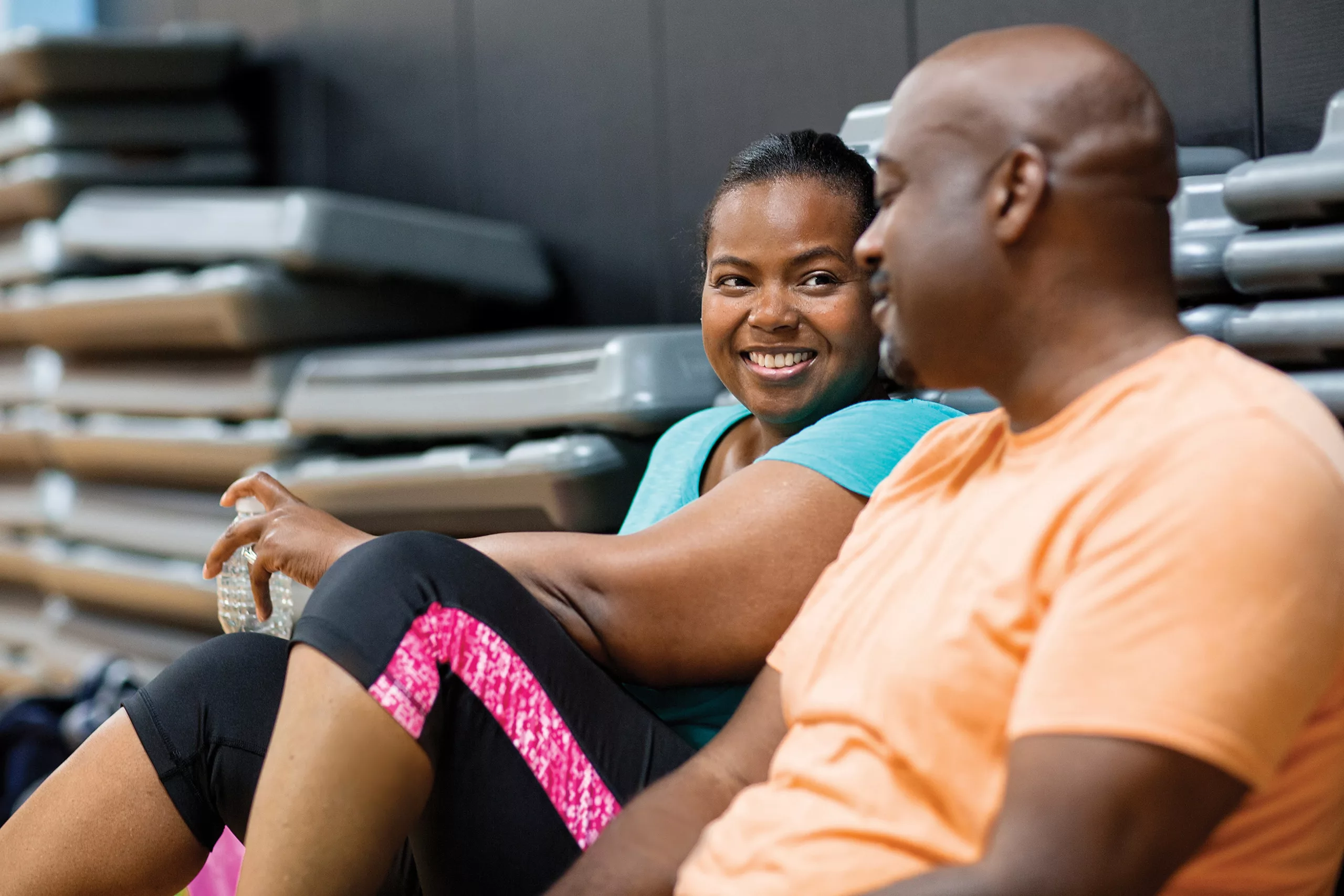 Two people sitting and talking, smiling after a workout, with exercise steps in the background.